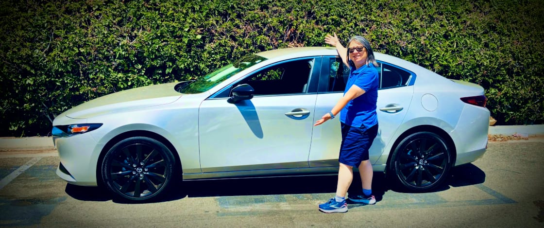 A happy woman stands, spokesmodel-style, next to a beautiful 2025 Mazda3 sedan.